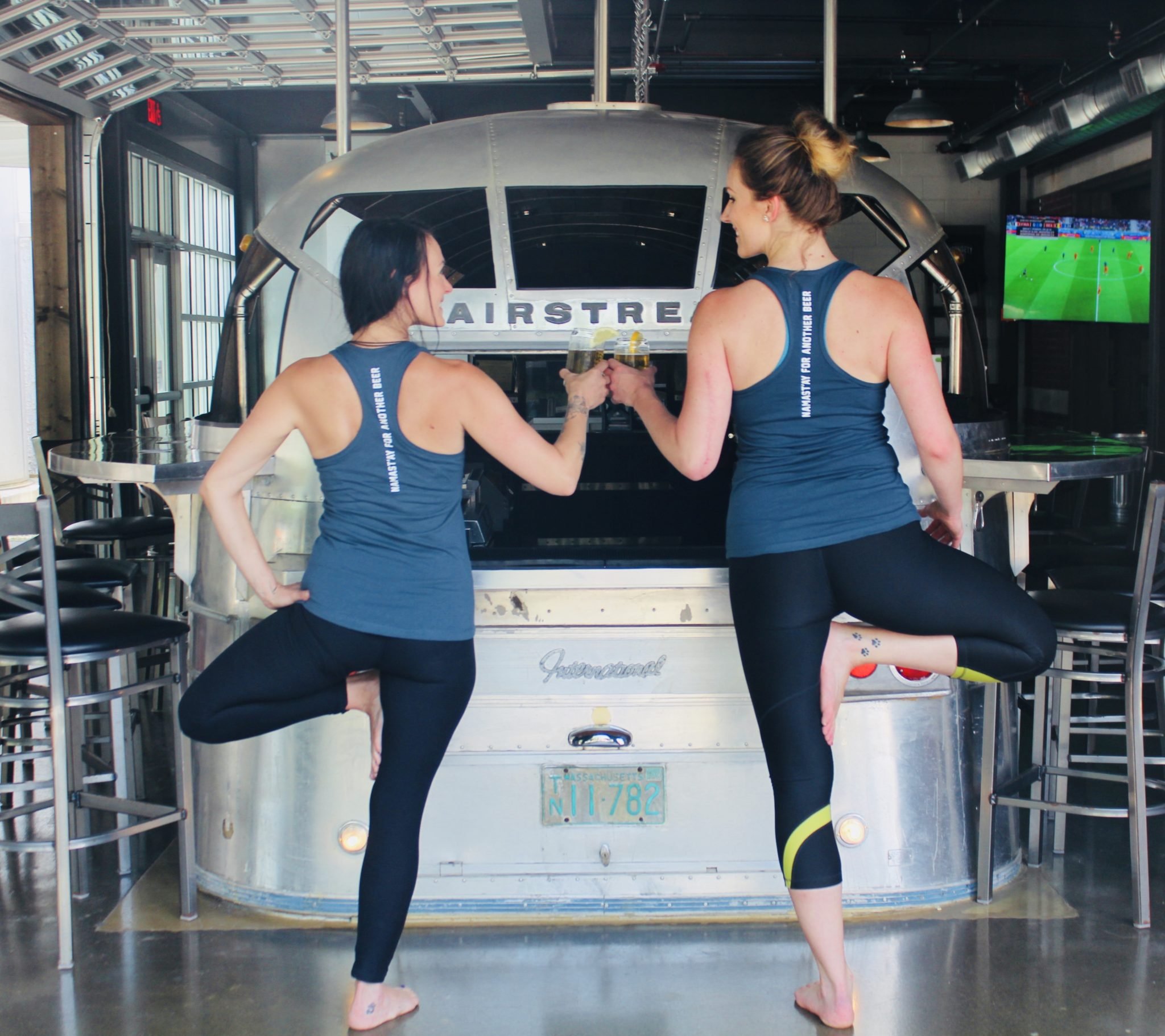 Two women in athletic wear doing a yoga pose while holding drinks in front of a vintage Airstream trailer bar.
